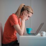 A woman sits indoors at a table, looking concerned while using a laptop with headphones and a coffee mug nearby.